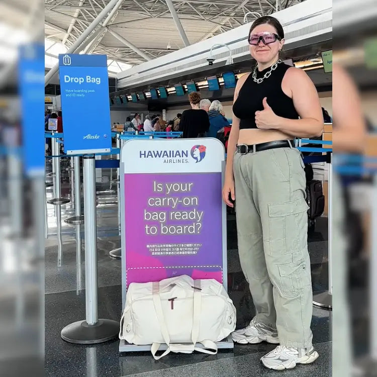 Person standing at an airport with a Hawaiian Airlines sign and a white bag.