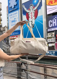 Person holding a beige duffel bag in an urban setting with advertisements in the background