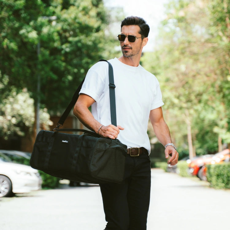Man in white t-shirt and sunglasses carrying a black duffel bag outdoors.