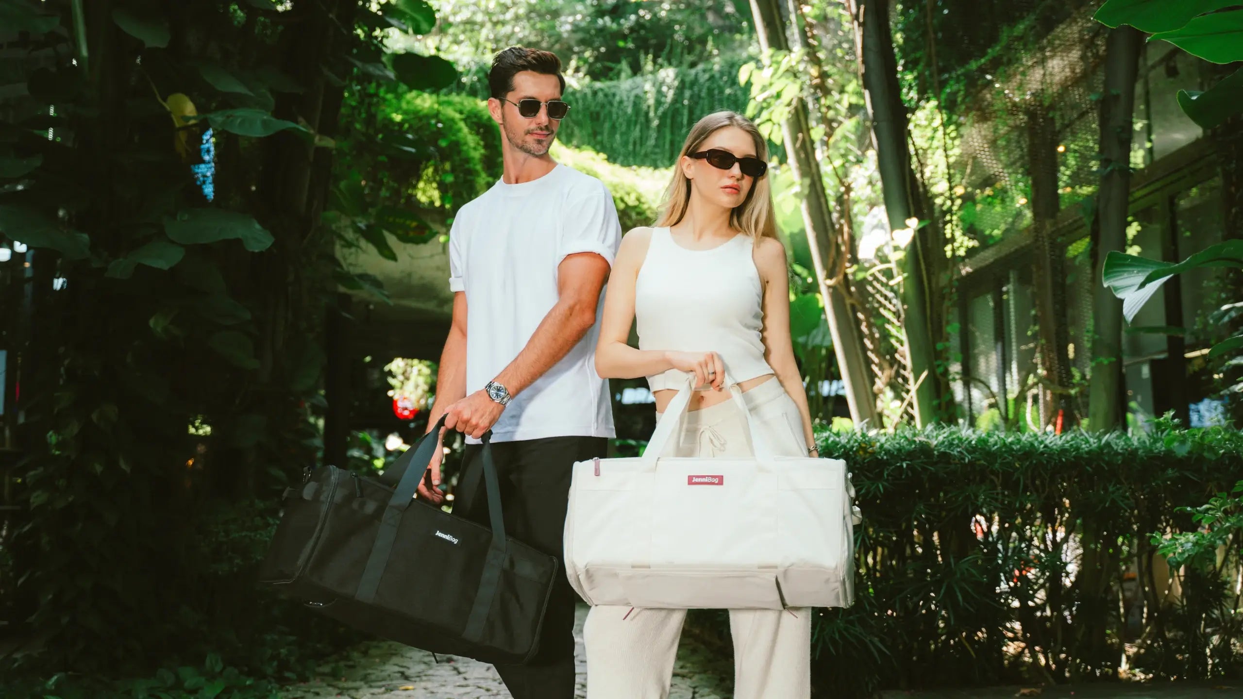 Two people walking outdoors with bags, surrounded by greenery