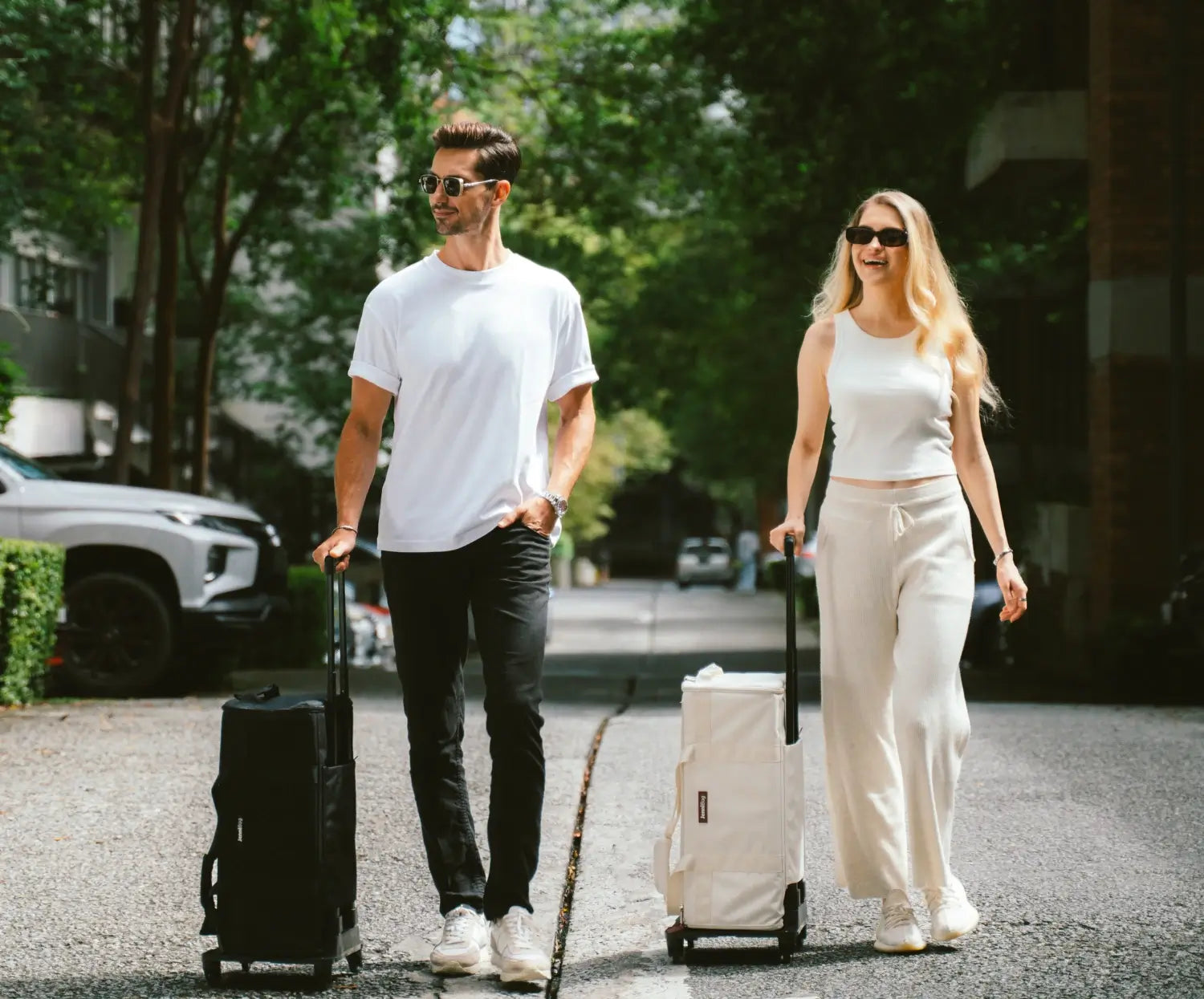 Man and woman walking with suitcases on a path surrounded by trees.