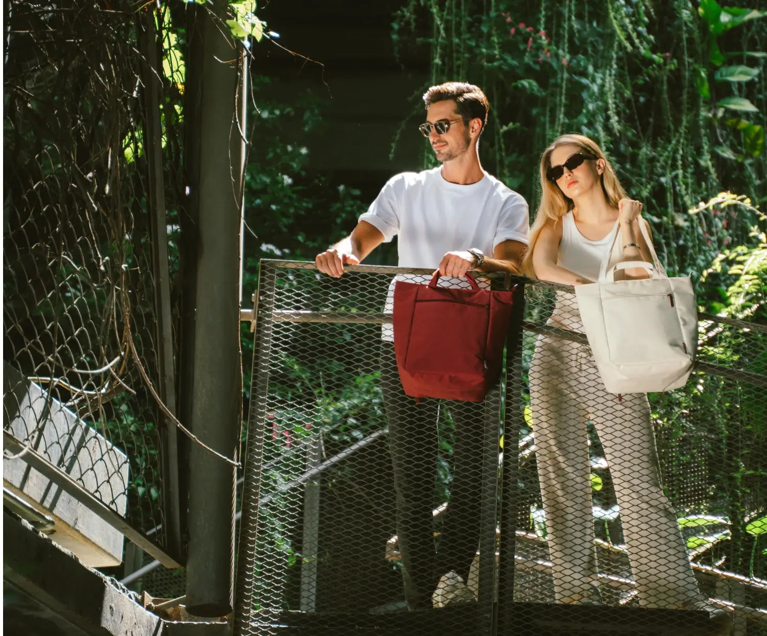 Two people walking on a bridge in a forested area, each carrying a bag.
