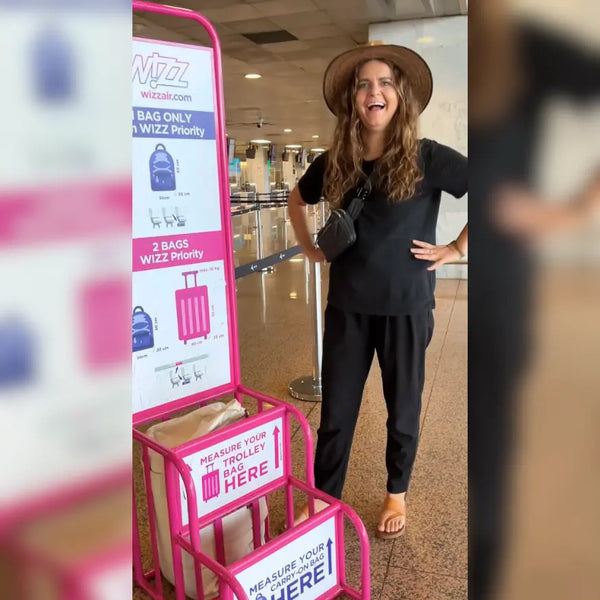 Traveler laughing beside Wizz Air’s pink “Measure your trolley here” sizer with a cream JenniBag inside
